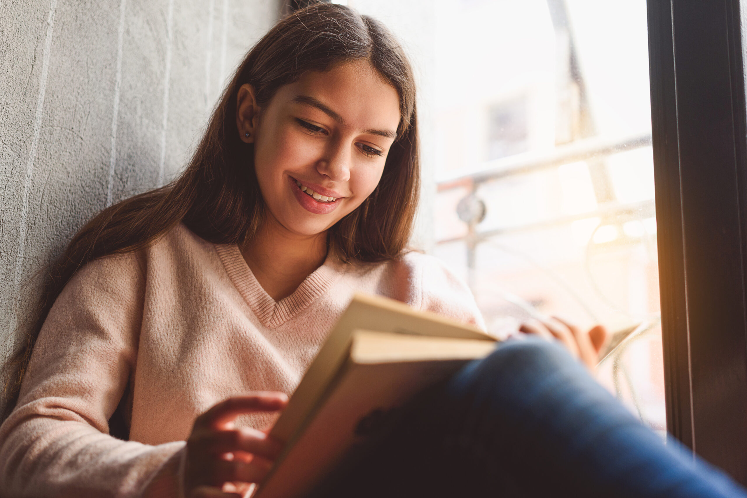 Smiling female teenager is on a window sill reading a book