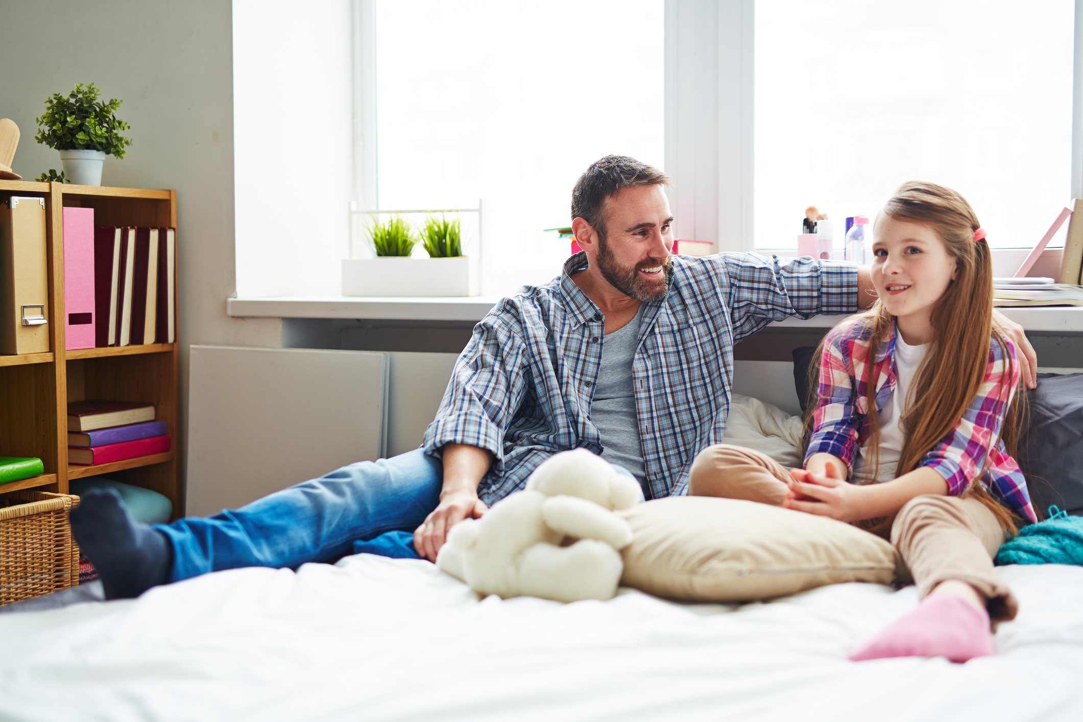 Teenage daughter in checked shirt sitting on bed next to her father and telling him about problems, he listening to her attentively and giving advice